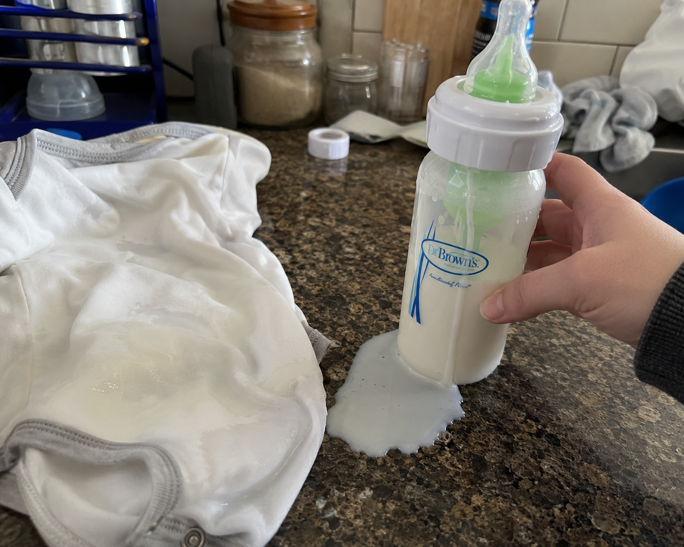 Milk leaking from a plastic bottle onto a baby outfit and kitchen counter